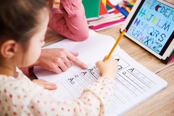 Teacher helping young child write the letter A on a worksheet in a notebook.