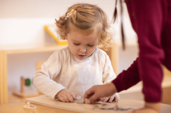 Girl playing with wooden puzzle with help from a teacher at childcare center.