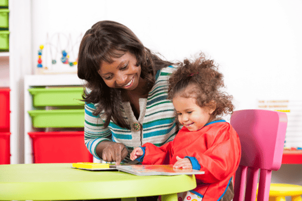 Teacher sits with young child at a table, reading a picture book