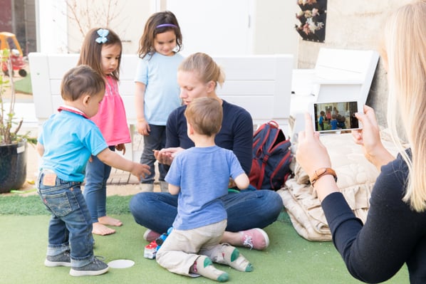 A teacher sits on the ground holding out her hand to show a group of four toddlers around her. In the foreground, another woman uses her phone to take a picture of the scene in front of her.