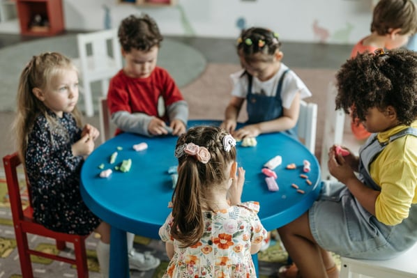 Five children seated around a blue round table engaged in small parts play.