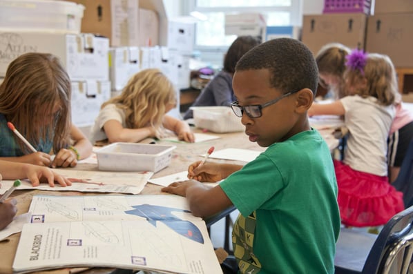 Young boy in a green t-shirt and glasses looks at an open textbook and takes notes on a notepad.