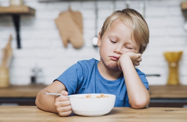 Little boy sitting at a counter, resting his head on his hand, looking down at a bowl of untouched cereal in front of him.