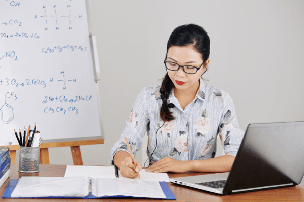 female teacher sitting at desk lesson planning, writing in a notebook with her laptop open