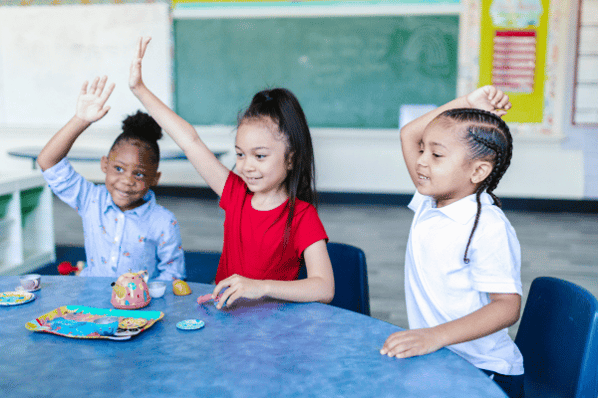 Three young children sitting at a table with their hands raised 