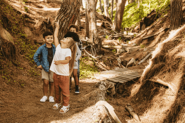 Children on a nature walk in the woods. One young girl in the front of the group is pointing her finger and looking up toward the sky.