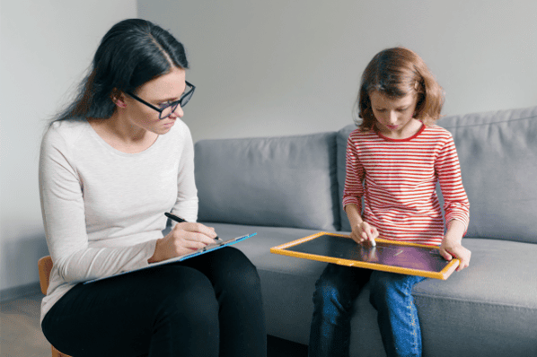 Teacher with clipboard observing young child drawing on a tablet.