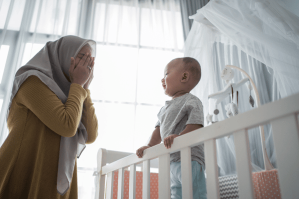 Woman playing peek-a-boo with baby standing up in a crib.