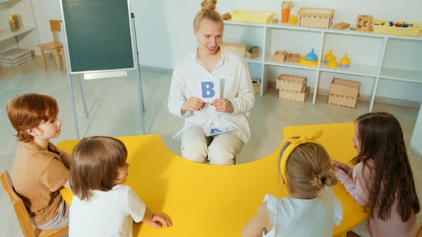 Preschool teacher showing a card with the letter 'B' to students seated at a low table.