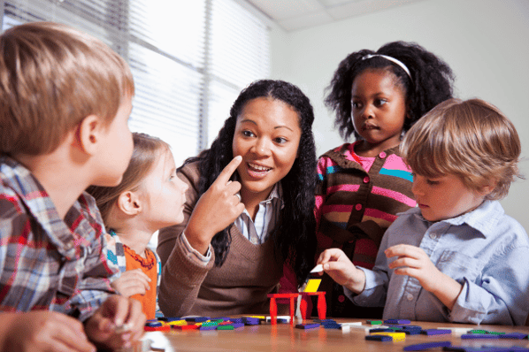 A group of preschool children working with legos at a table. Their teacher is sitting in the middle of the table explaining a concept to them. The children are paying attention to their teacher's instruction.