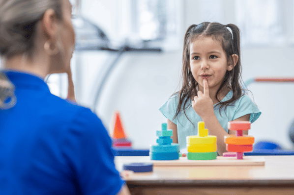 Young girl at speech therapy, sitting at desk touching her pointer finger to her chin. 