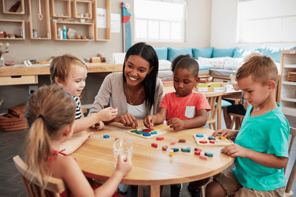 Female teacher sitting at a round wooden table with four young children playing with wooden puzzles.