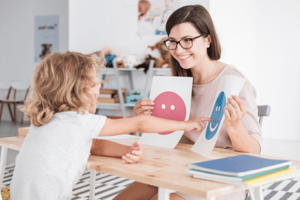 A female counselor holding pictures of a red sad face and a happy blue face during a meeting with a young patient. The patient is pointing to the blue happy face.