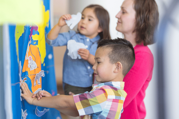 two young children glue animal photos onto a map of the world hanging on a wall while their teacher watches